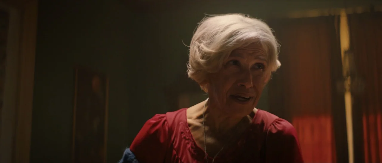 The Front Room (2024) movie still: closeup — A dimly lit interior room features dark walls and soft lighting. An older woman with short light hair…