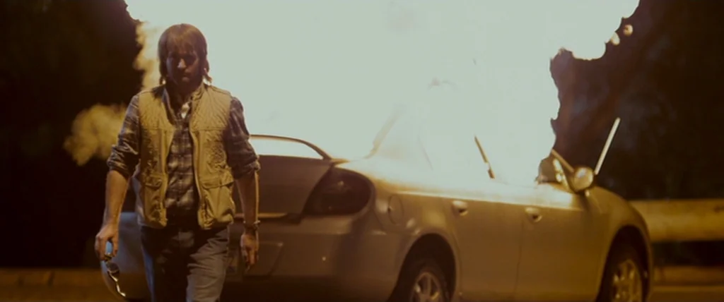MacGruber (2010) movie still: fullbody — A man walks away from a car engulfed in flames on an empty road at night. Bright orange fire consumes the…
