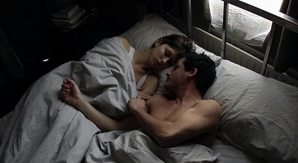 9 Songs (2004) movie still: medium — A couple lies in a bed with grey sheets near a window. They are resting together side by side with eyes closed.
