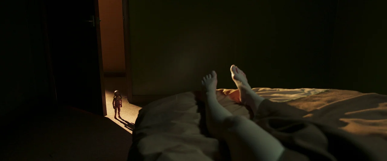 Stopmotion (2024) movie still: wide — The interior of a dimly lit room seen from the perspective of a reclining person. A small doll stands in the…
