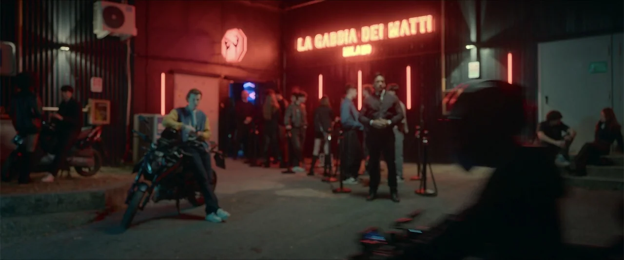 Love Me Love Me (2026) movie still: wide — A dark urban exterior features a glowing red neon sign above a club entrance. People wait in a line while…