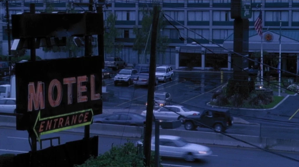 Broken Flowers movie still: wide — A motel parking lot sits adjacent to a dark road at night with various cars parked. A large illuminated sign for…