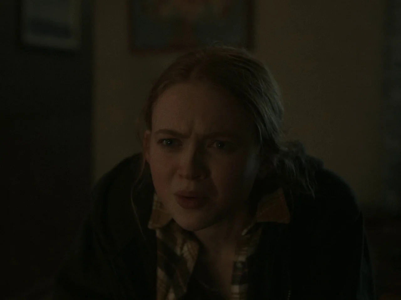 The Whale (2022) movie still: closeup — A dimly lit indoor space with indistinct dark walls in the background. A young woman with red hair looks…