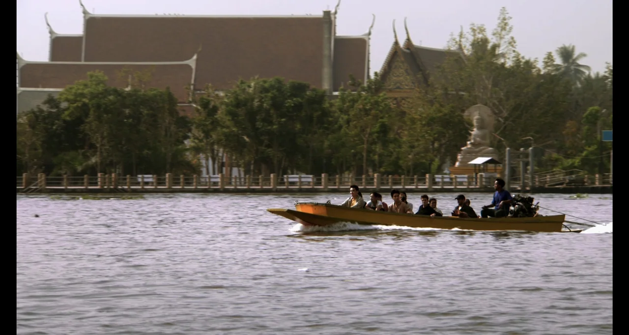 Ninja: Shadow of a Tear (2013) movie still: wide — A longtail boat filled with passengers navigates a wide river under a clear sky. A traditional…