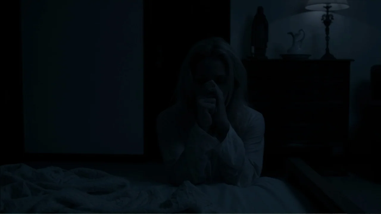 The Grotto (2022) movie still: medium — A dark bedroom at night features a bed and a dresser with a lamp. A woman kneels on the bed with her hands…