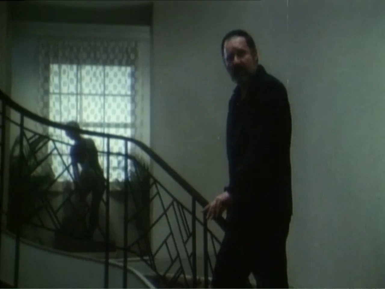 Curtains (1995) movie still: medium — A man walks down a staircase with a black geometric metal railing. A window with a patterned curtain stands in…