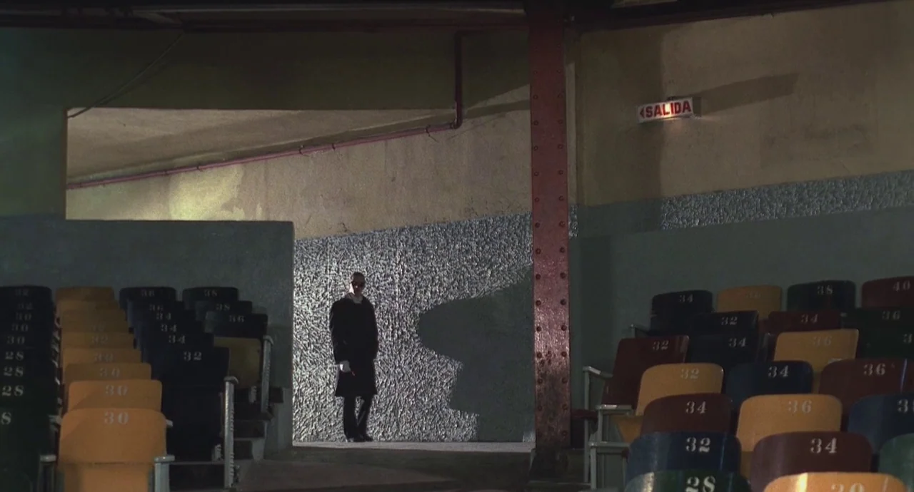 Santa Sangre (1989) movie still: wide — An empty, dimly lit concrete corridor sits between rows of weathered stadium seats. A solitary man in a dark…