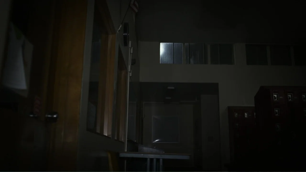 The Gallows (2015) movie still: wide — An empty school hallway features rows of dark metal lockers and tall glass windows. Dim light filters through…