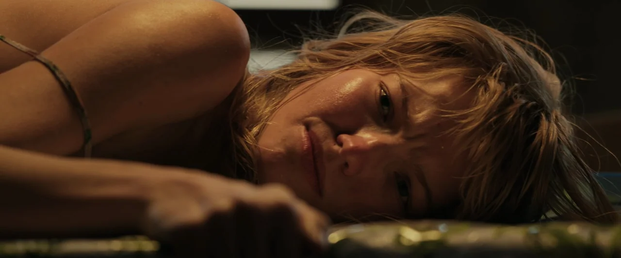 Anna (2019) movie still: closeup — A woman lies down on a flat surface in a dimly lit indoor space. She rests her chin near the foreground while…
