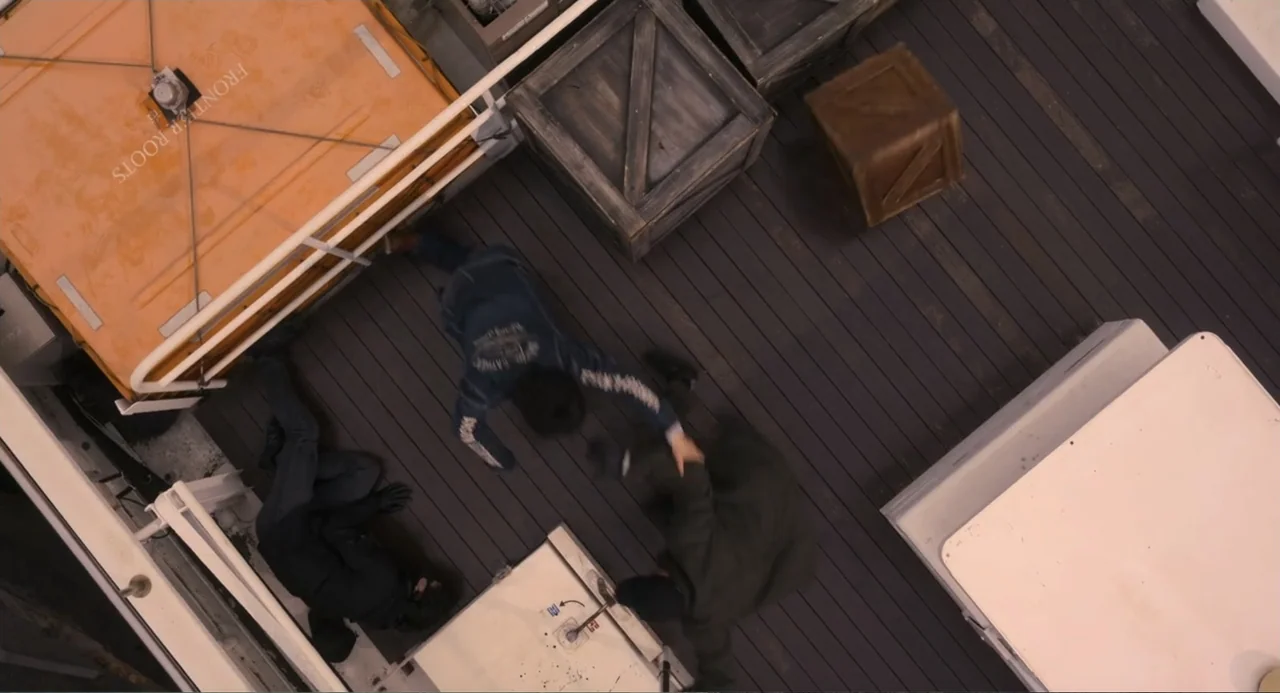 Knuckle Girl (2023) movie still: wide — A wooden ship deck is cluttered with wooden crates and an orange life raft. Three men engage in a physical…