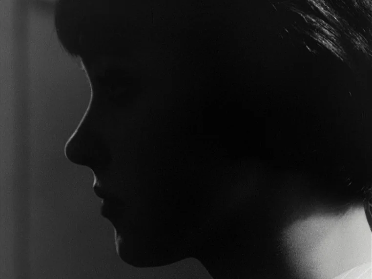 Vivre Sa Vie (1962) movie still: closeup — A dimly lit room provides a black backdrop for a side profile shot. A woman stands with her face…