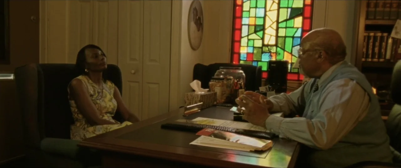 Sermon on the Mount (2019) movie still: medium — An interior office space features a dark wooden desk and a colorful stained glass window. A man…