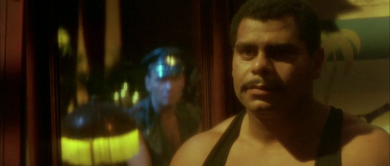 Querelle (1982) movie still: closeup — An interior shot shows a man in a dark tank top standing before a mirror. A reflection of a person in a cap…