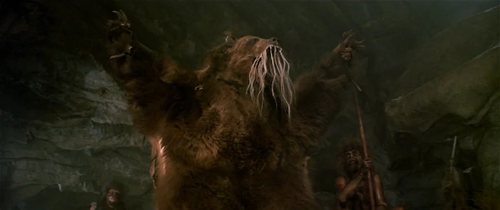 Clan of the Cave Bear (1986) movie still: medium — A dark, rocky cave interior is dimly lit by flickering firelight. A man wears a large bear skin…