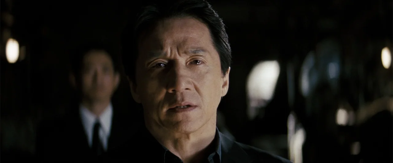 Rush Hour 3 (2007) movie still: closeup — A man stands in a dark interior space with an indistinct figure blurred behind him. The man looks forward…