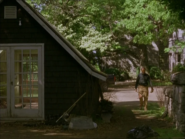 Last Days (2005) movie still: wide — A dark wooden shed with double glass doors sits beside a gravel path. A man walks toward the camera carrying a…