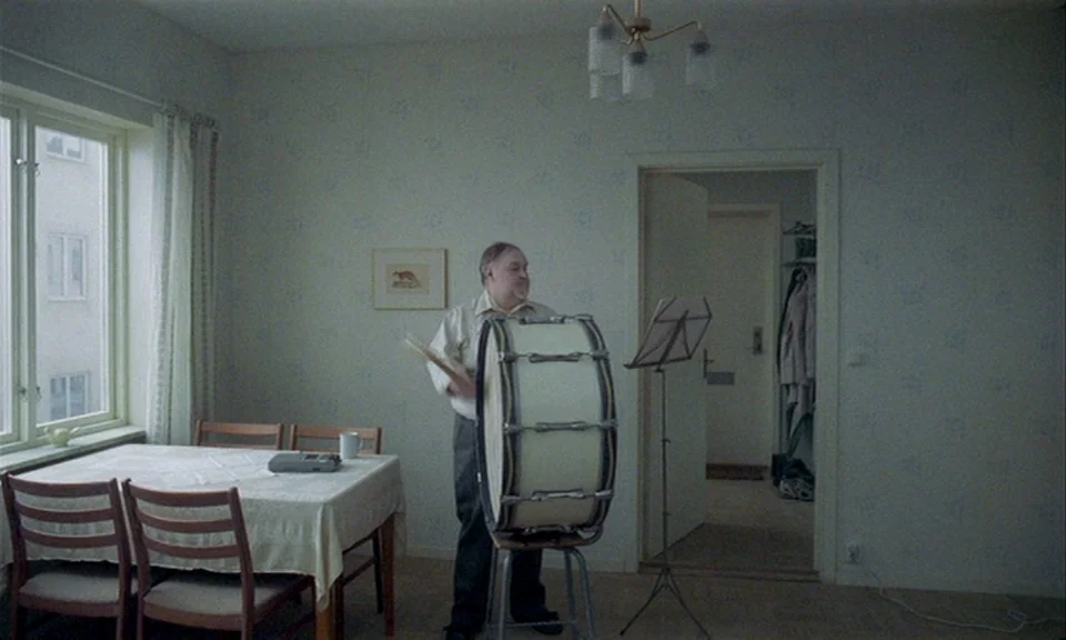 You, The Living (2007) movie still: fullbody — A sparsely furnished room contains a dining table and a man with a drum. The man stands next to a…