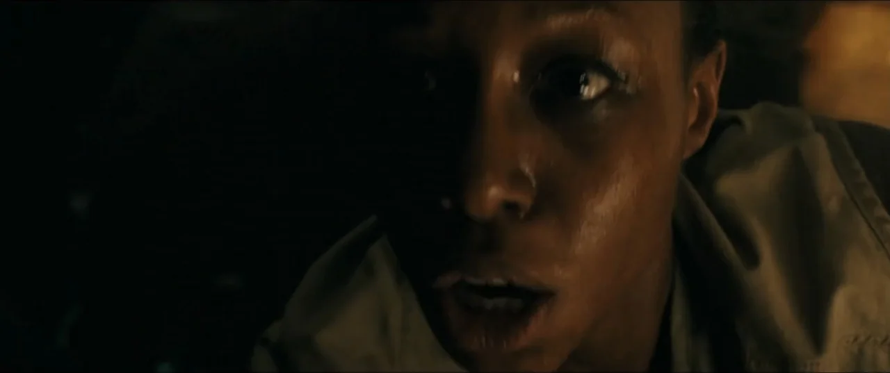 mother! (2017) movie still: closeup — A dark, obscured interior setting with minimal light visible. A woman with a beige vest stares forward with an…