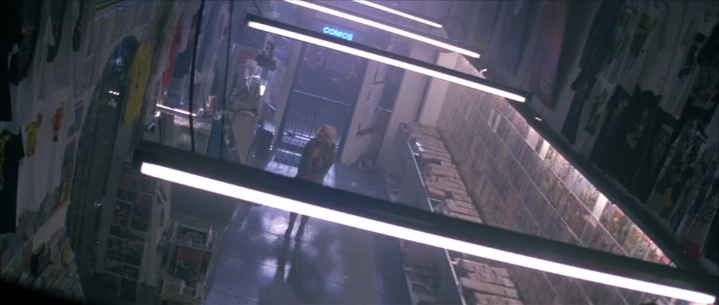 True Romance (1993) movie still: wide — A cluttered comic book shop interior with long shelves and hanging apparel. A lone woman stands in the…
