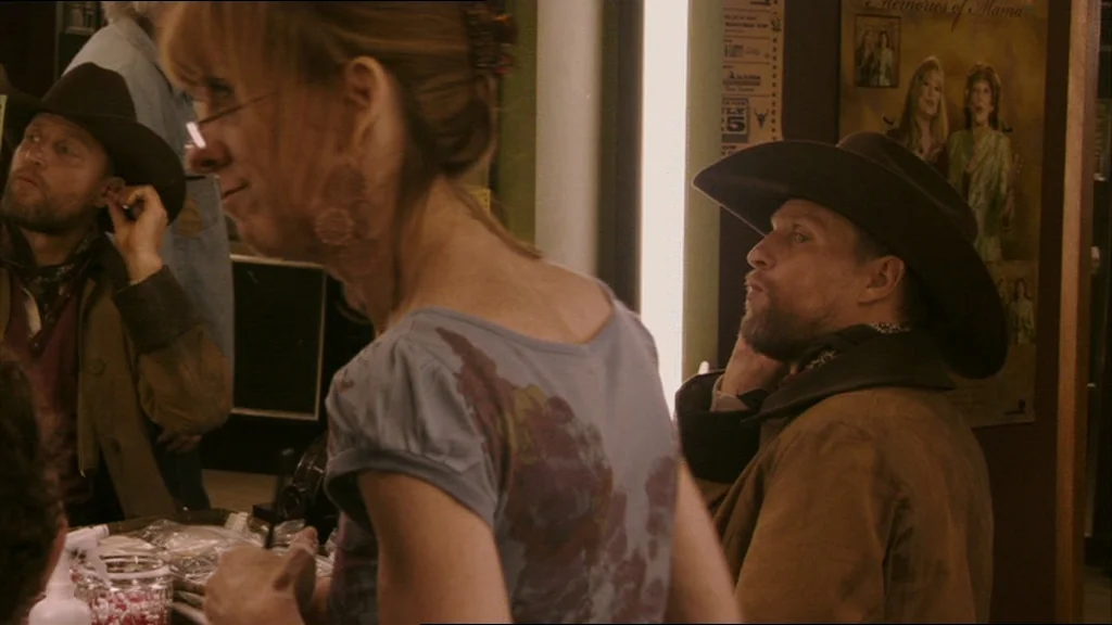 A Prairie Home Companion movie still: medium — A cluttered interior with posters and professional hair supplies visible on a vanity. A woman stands…
