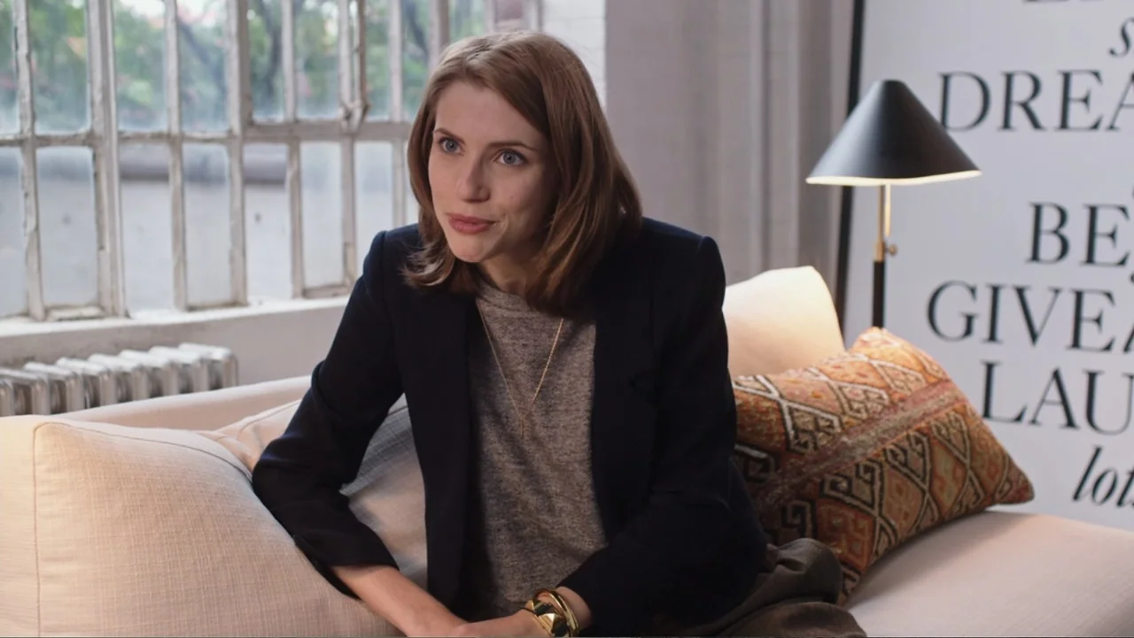 The Intern (2015) movie still: medium — A light-filled living room features a white sofa, a framed poster, and grid windows. A young woman sits on…