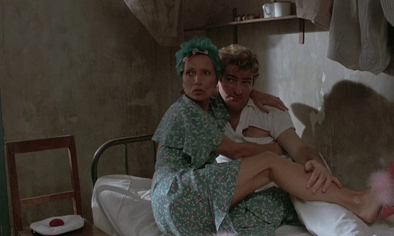 Coup De Torchon (1981) movie still: medium — A cramped, dimly lit room with peeling beige walls and basic furniture. A woman in a patterned dress…