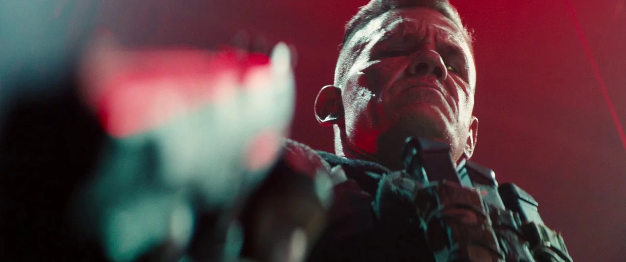 Deadpool 2 (2018) movie still: closeup — A dark industrial environment is washed in intense red light. A muscular man with scars and a cybernetic…