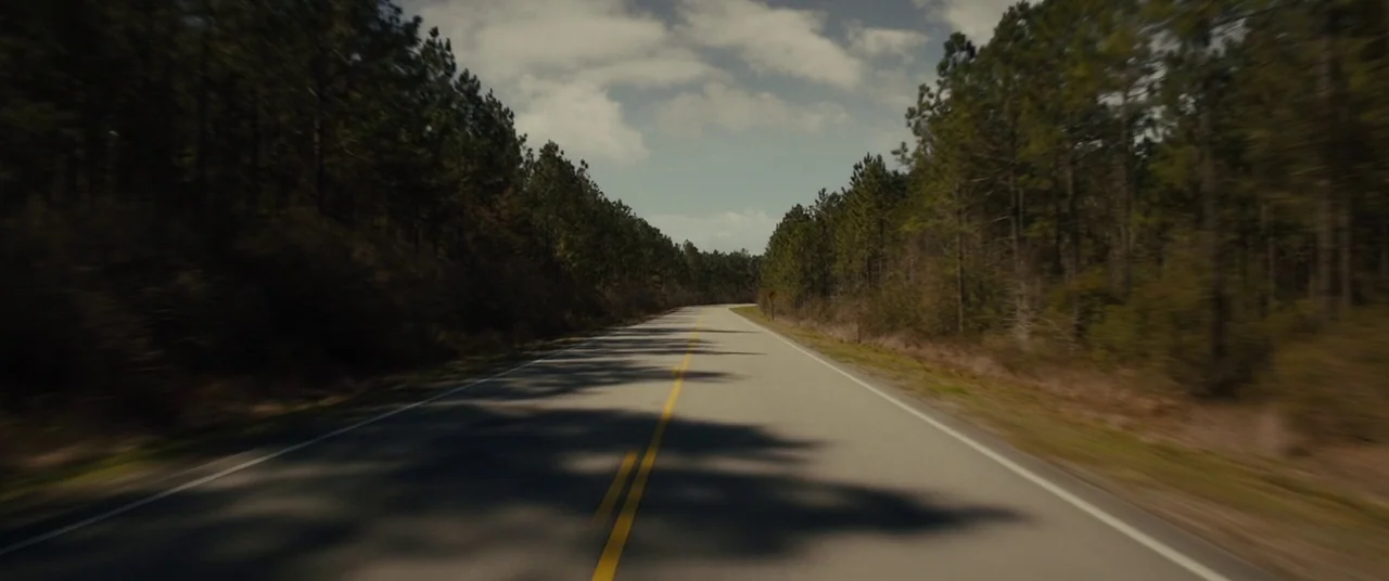 Midnight Special (2016) movie still: establishing — A long asphalt road cuts through a dense pine forest under a cloudy sky. The camera moves along…