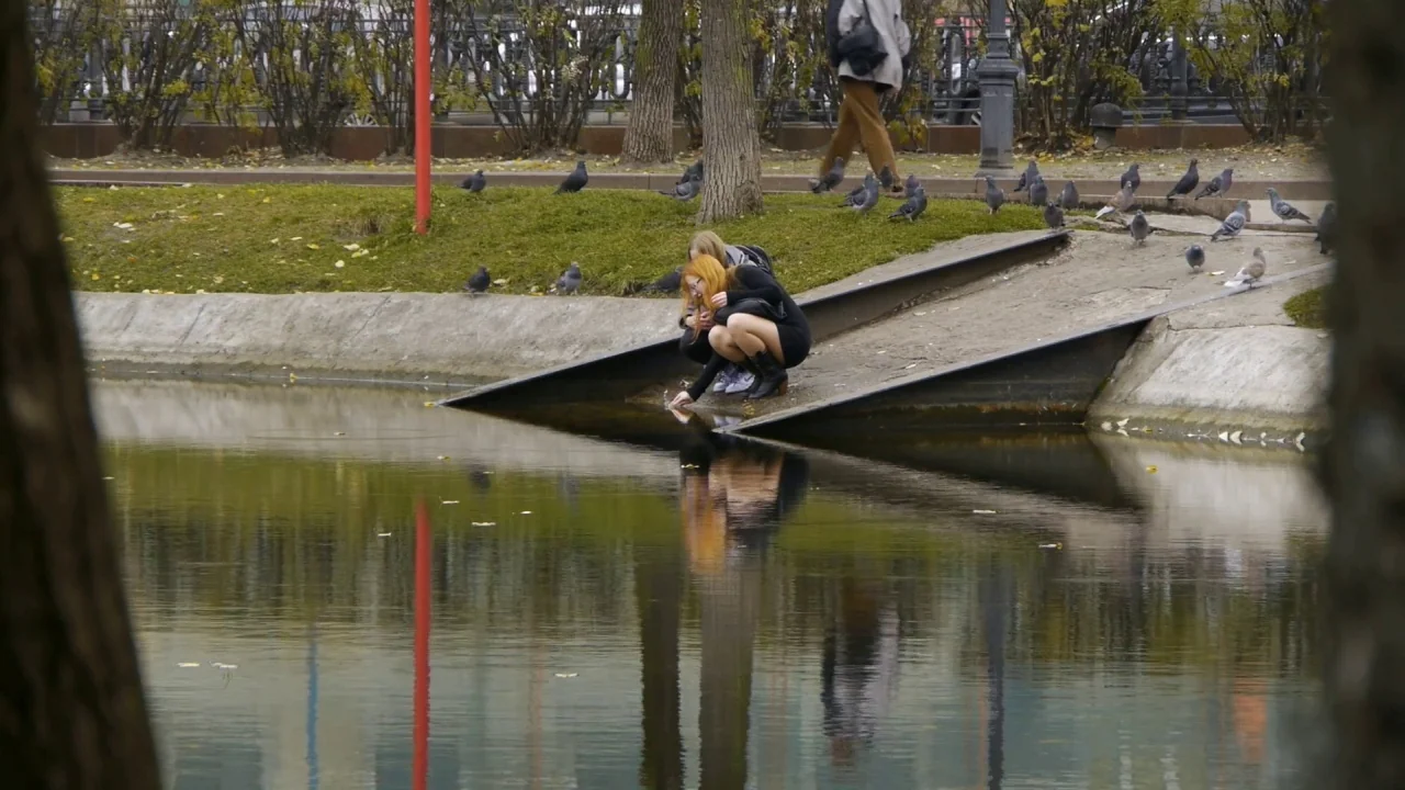 Counting (2015) movie still: wide — A park pond features a slanted concrete ramp leading into the water. Two women crouch at the water's edge with…
