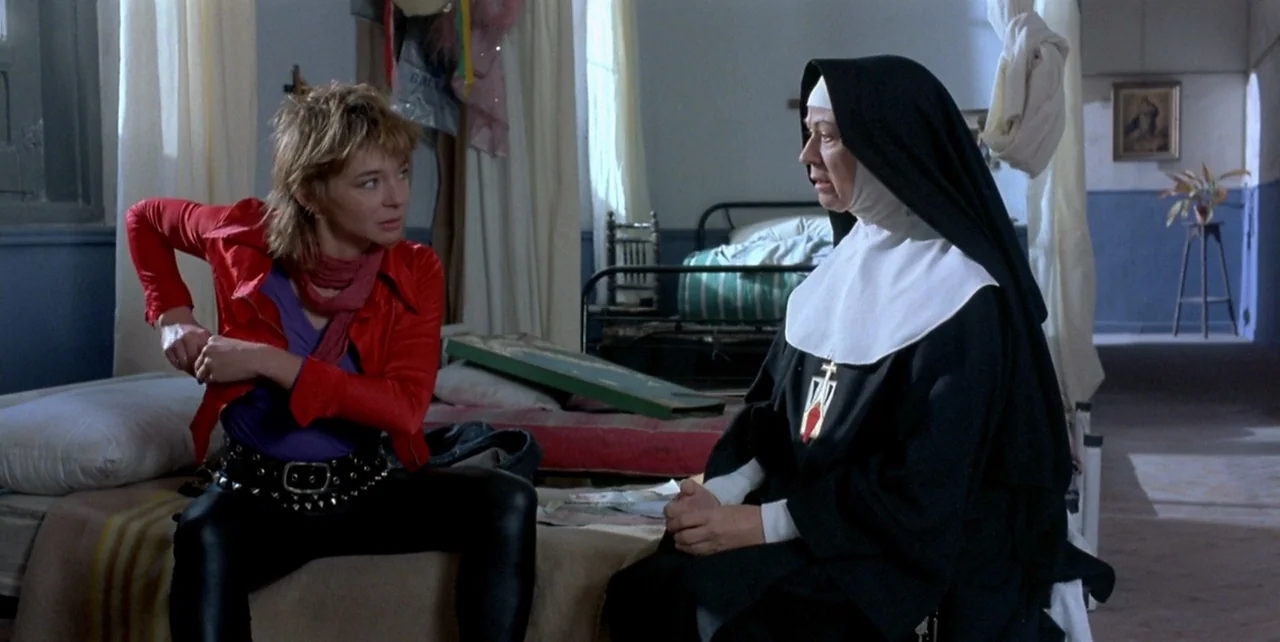 Dark Habits (1983) movie still: medium — A spartan dormitory features simple metal bed frames and light-colored curtains against blue walls. A young…
