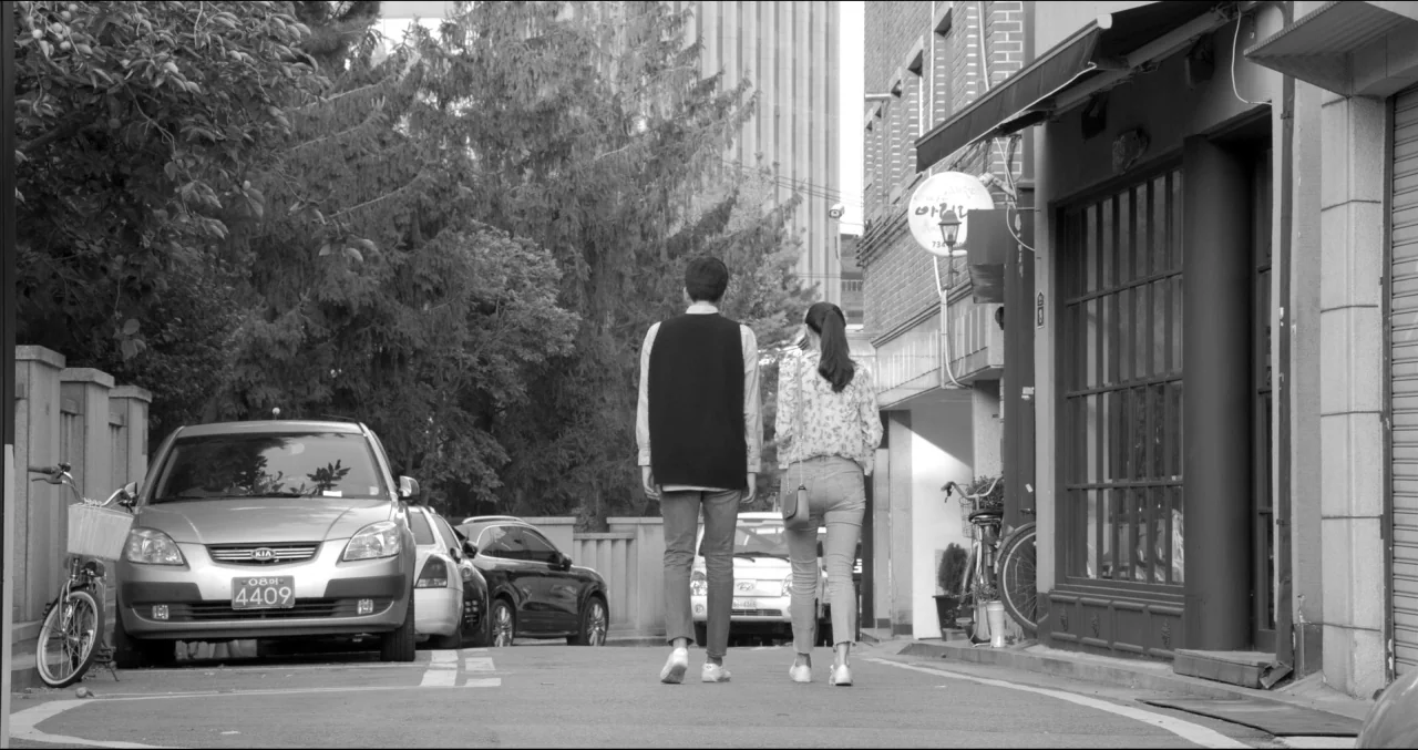 Grass (2018) movie still: fullbody — A narrow paved urban road is lined with parked cars and storefronts on one side. A young man and woman walk…