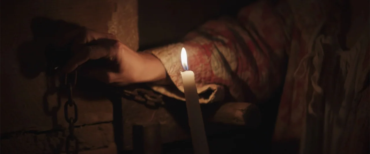 The Wind (2005) movie still: closeup — A dark stone or wood interior with minimal illumination from a handheld candle. A person's hand reaches…