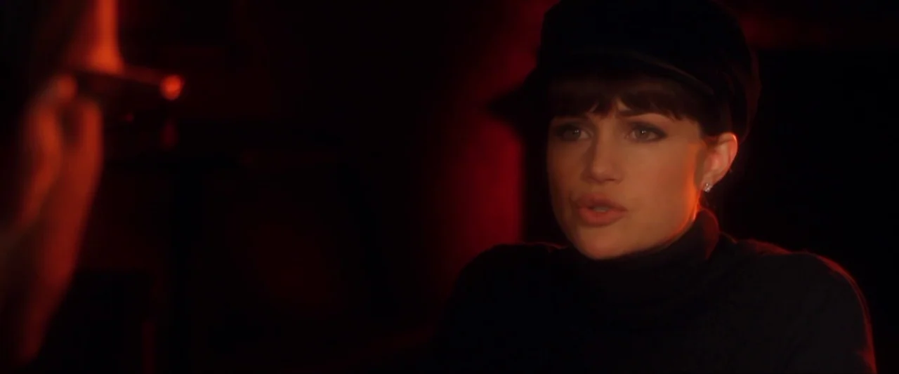 Girl Walks Into a Bar (2011) movie still: closeup — A dark, dimly lit environment is bathed in a pervasive red light. A young woman with a black cap…