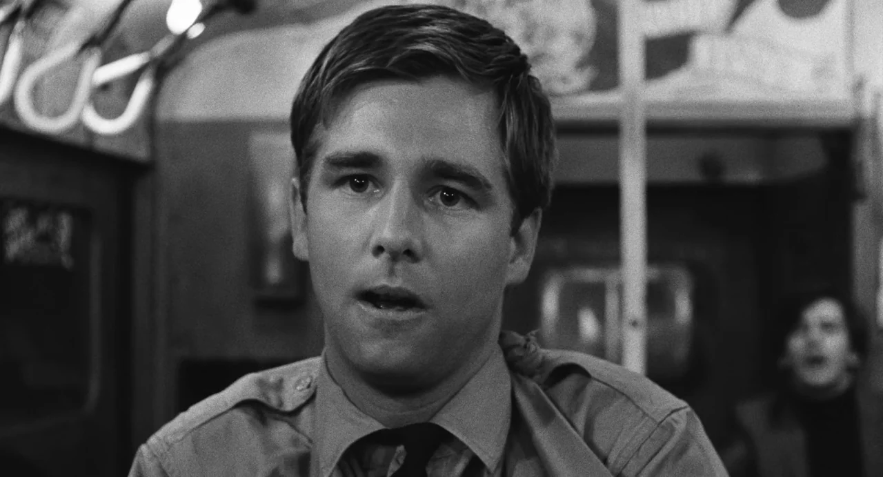 The Incident (1967) movie still: closeup — A man sits inside a public vehicle with interior rails visible. He gazes forward with a neutral…
