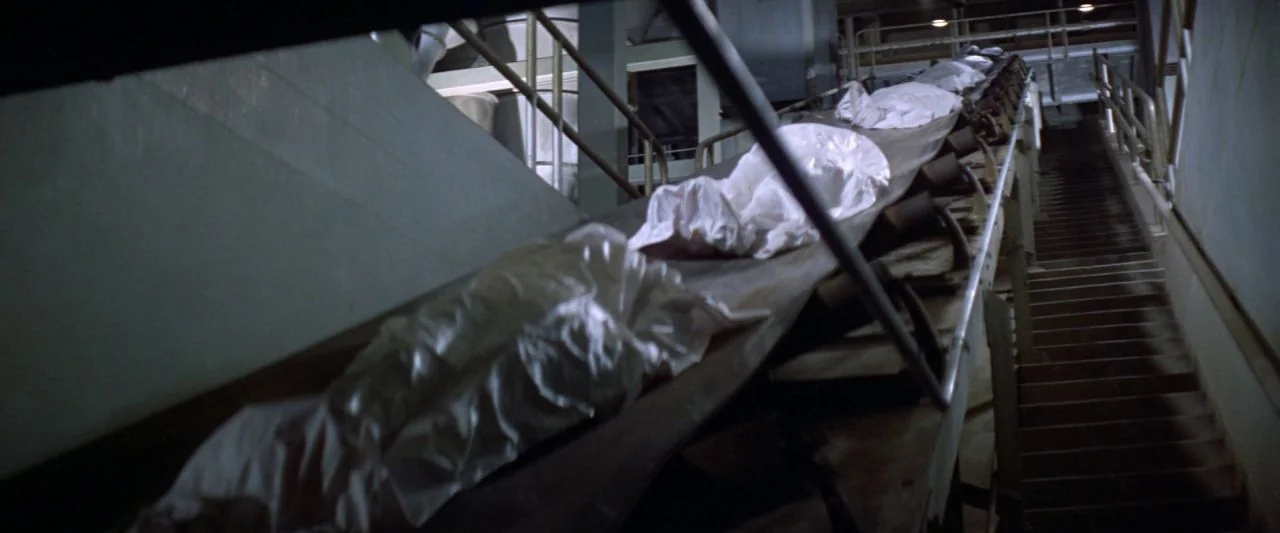 Soylent Green (1973) movie still: wide — A long mechanized conveyor belt system is housed within an industrial processing facility. Multiple bundles…