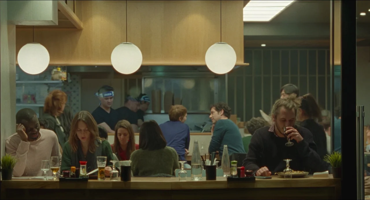 One Fine Morning (2022) movie still: wide — A modern restaurant features a long wooden bar counter with several patrons eating dinner. Staff wearing…