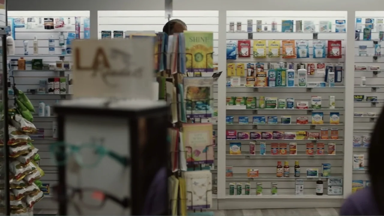 The Cleaner (2021) movie still: medium — A well-lit drugstore aisle features white slatwall shelving filled with various pharmaceutical products. A…