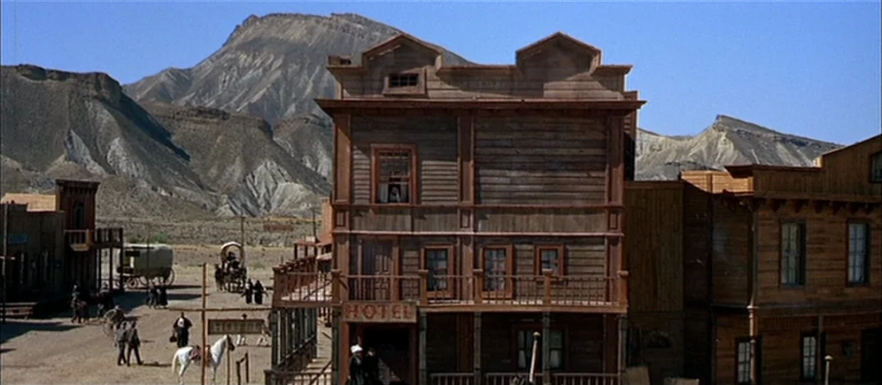 For A Few Dollars More (1965) movie still: wide — A wooden hotel stands in a dusty western frontier town with desert mountains behind. Several…