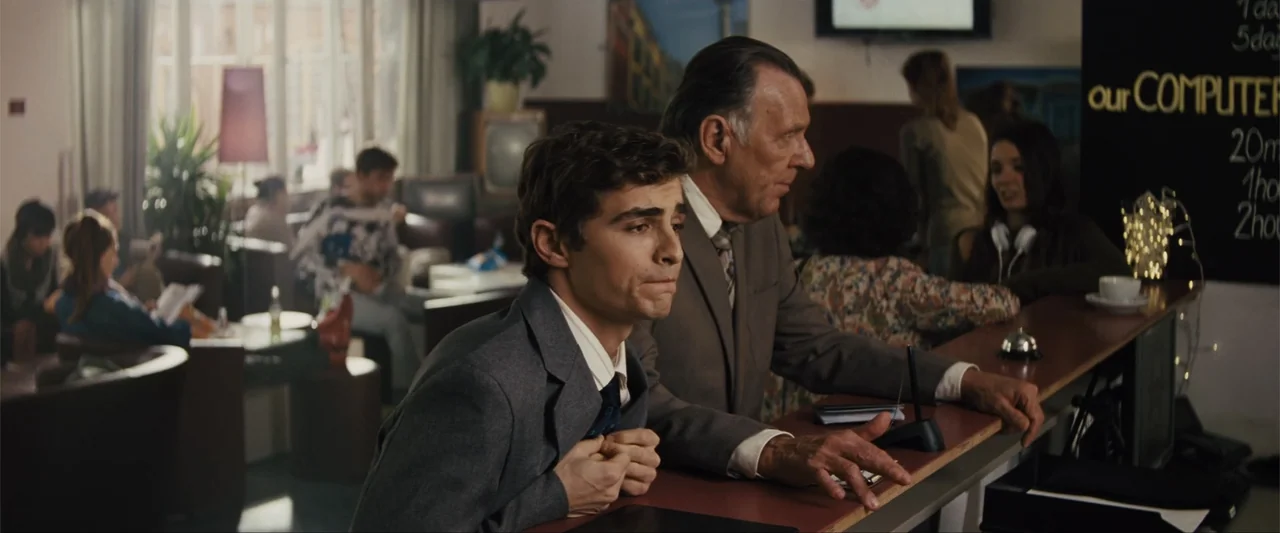 Unfinished Business (2015) movie still: medium — A hotel lobby with a reception desk, wood paneling, and various people in the background. A young…