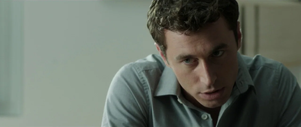 The Canyons (2013) movie still: medium — An interior space features a man looking down while seated in a brightly lit room. The focus is tight on…
