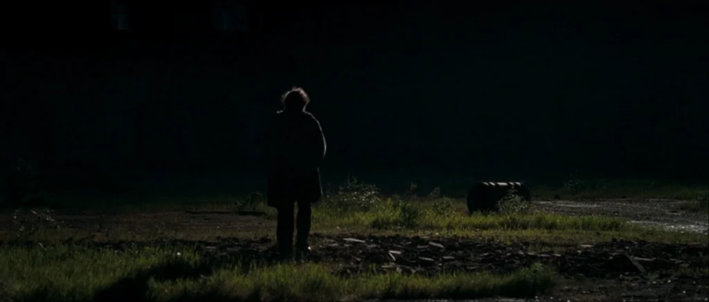 Happy-Go-Lucky (2008) movie still: wide — A dark, expansive outdoor area with sparse grass and uneven terrain. A single person stands in the center…