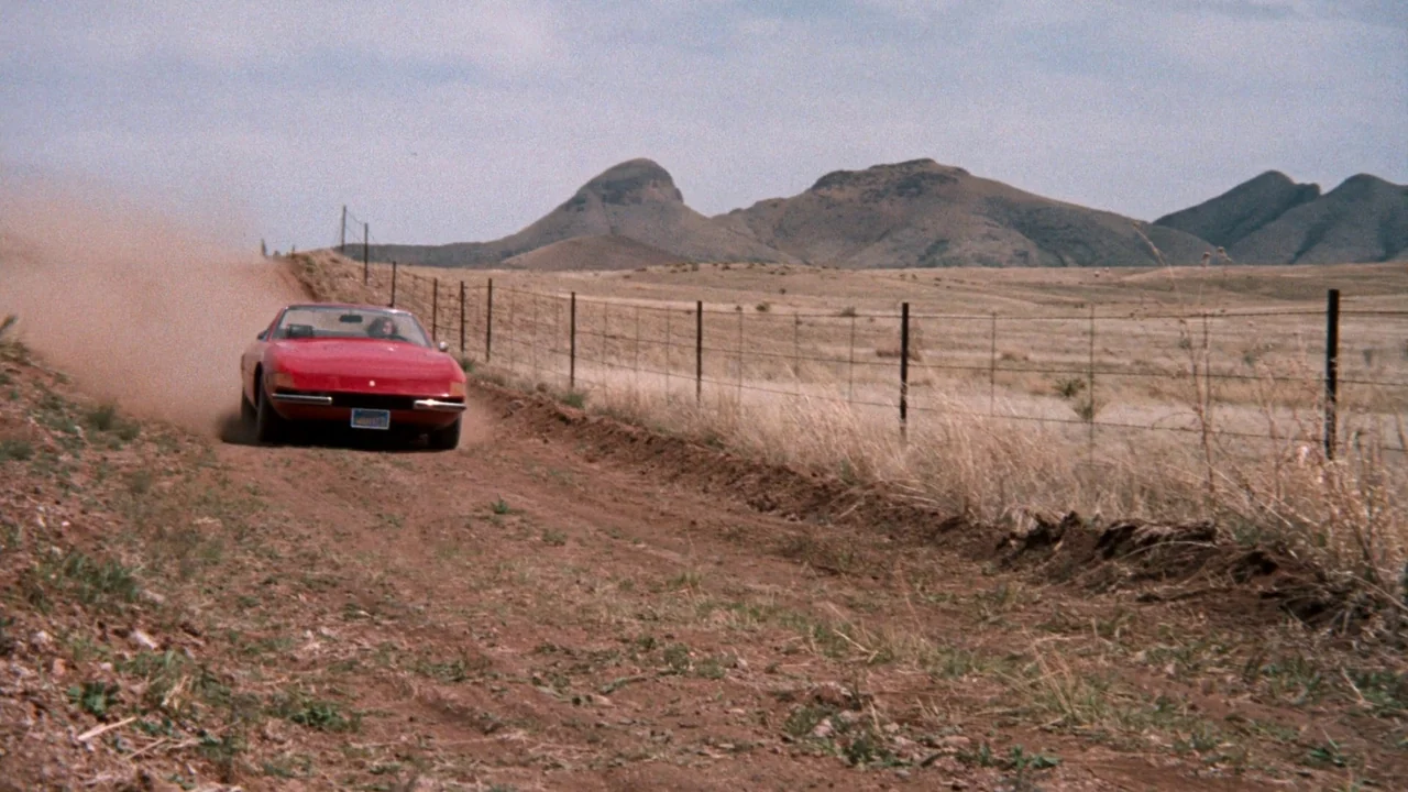 A Star Is Born (1976) movie still: wide — An arid, grassy expanse features a wire fence running parallel to a dirt road. A red convertible sports…
