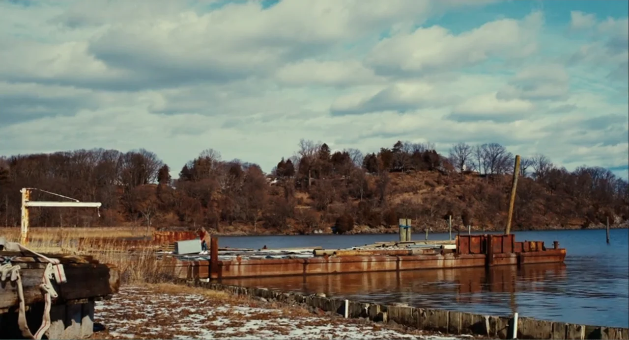 An Imperfect Murder (2017) movie still: wide — A rusted barge rests along a shoreline during late autumn or early winter. A woman stands on the…