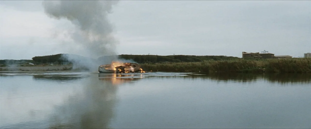 GOMORRAH (2023) movie still: wide — A burning wooden boat sits in a calm body of water near a reedy bank. A large plume of grey smoke rises from the…