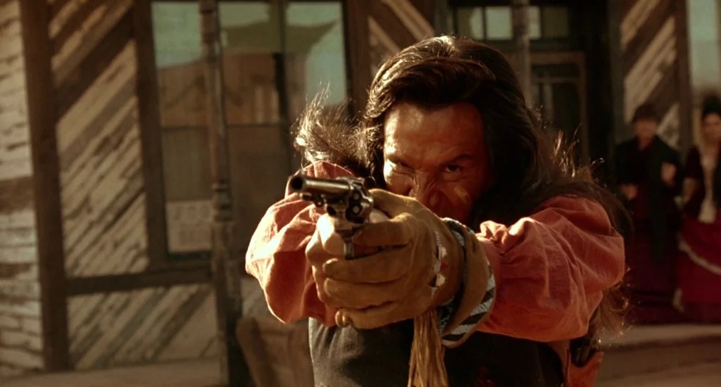 The Quick and the Dead (1995) movie still: medium — A wooden building with diagonal slats stands in a dusty frontier town. A man points a revolver…