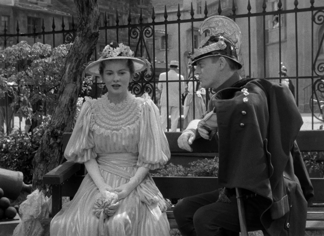 Letter From An Unknown Woman (1948) movie still: medium — A woman in a Victorian dress and a soldier in uniform sit on a bench. An iron gate and…