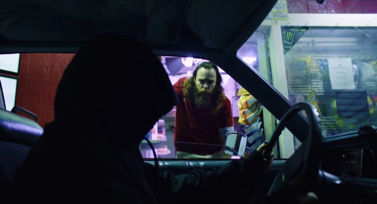 Like Me movie still: medium — A dark car interior faces the bright interior of a convenience store at night. A hooded figure sits in the driver seat…