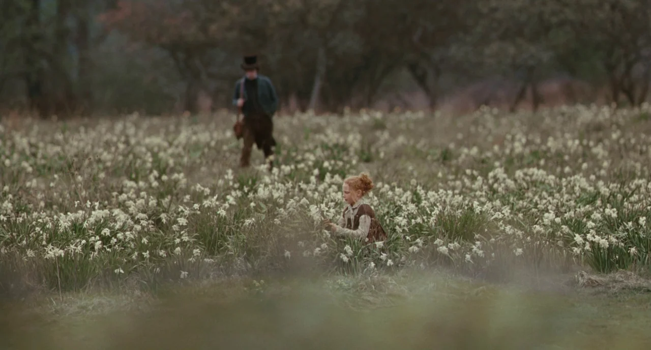 Bright Star (2009) movie still: wide — A field filled with white daffodils under a muted, overcast sky. A young girl kneels in the flowers while a…