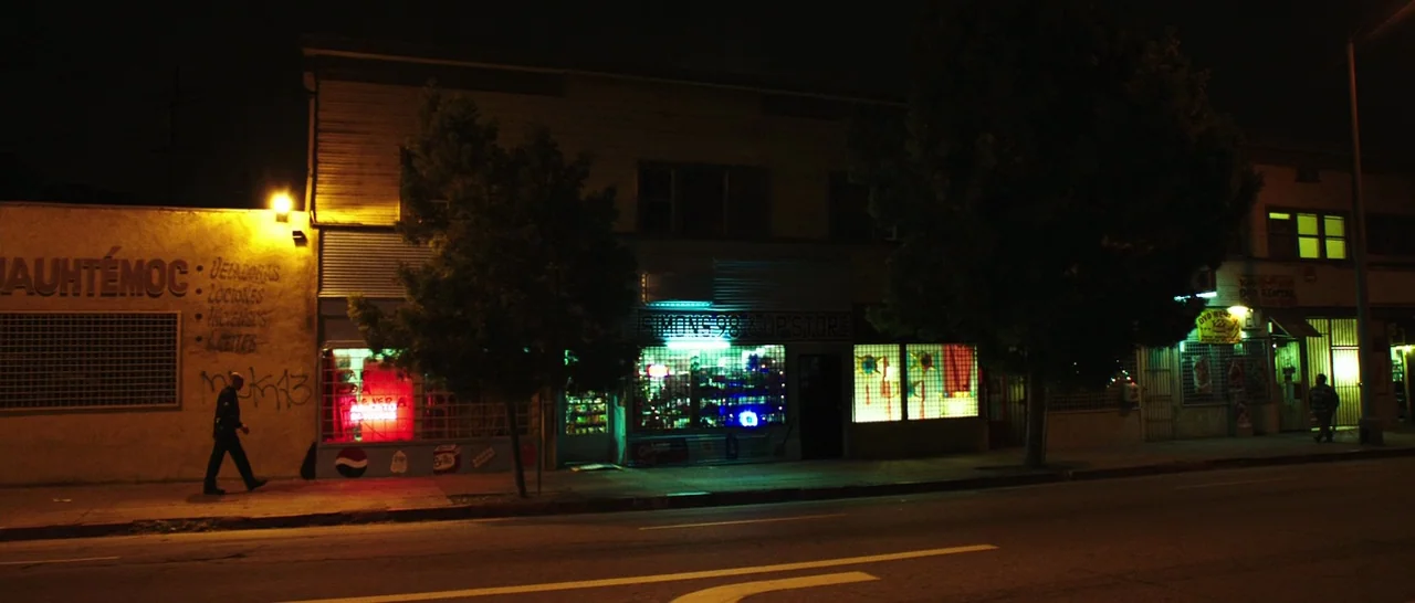 Rampart (2011) movie still: wide — A night view of a row of small commercial storefronts with illuminated signage. A single person walks along the…