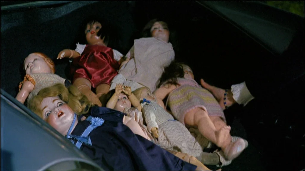 Chinese Roulette (1976) movie still: medium — Multiple vintage dolls lie scattered across the dark interior of an open car trunk. A human hand…